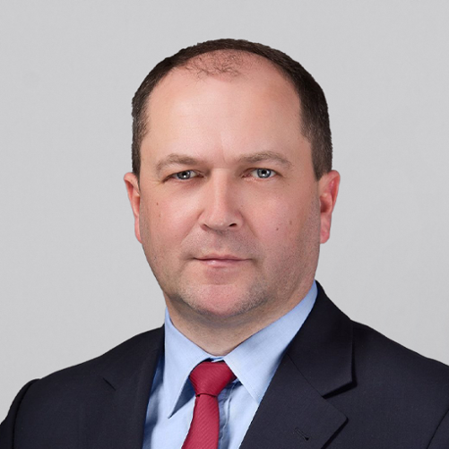 Na tle jasnoszarej ściany stoi wiceminister Kiepura w czarnym garniturze.