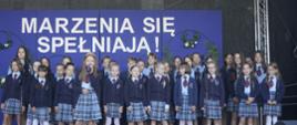 Na tle niebieskiej ściany z napisem Marzenia się spełniają stoi grupa ubranych na galowo dzieci.