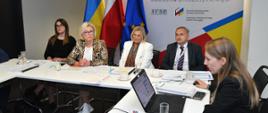 Przy białym stole siedzi kilka osób, z przodu kobieta w czarnym ubraniu patrzy w otwartego laptopa, za nimi flagi Polski, Ukrainy i UE, za nimi na ściance napis Polsko-Ukraińska Rada Wymiany Młodzieży.