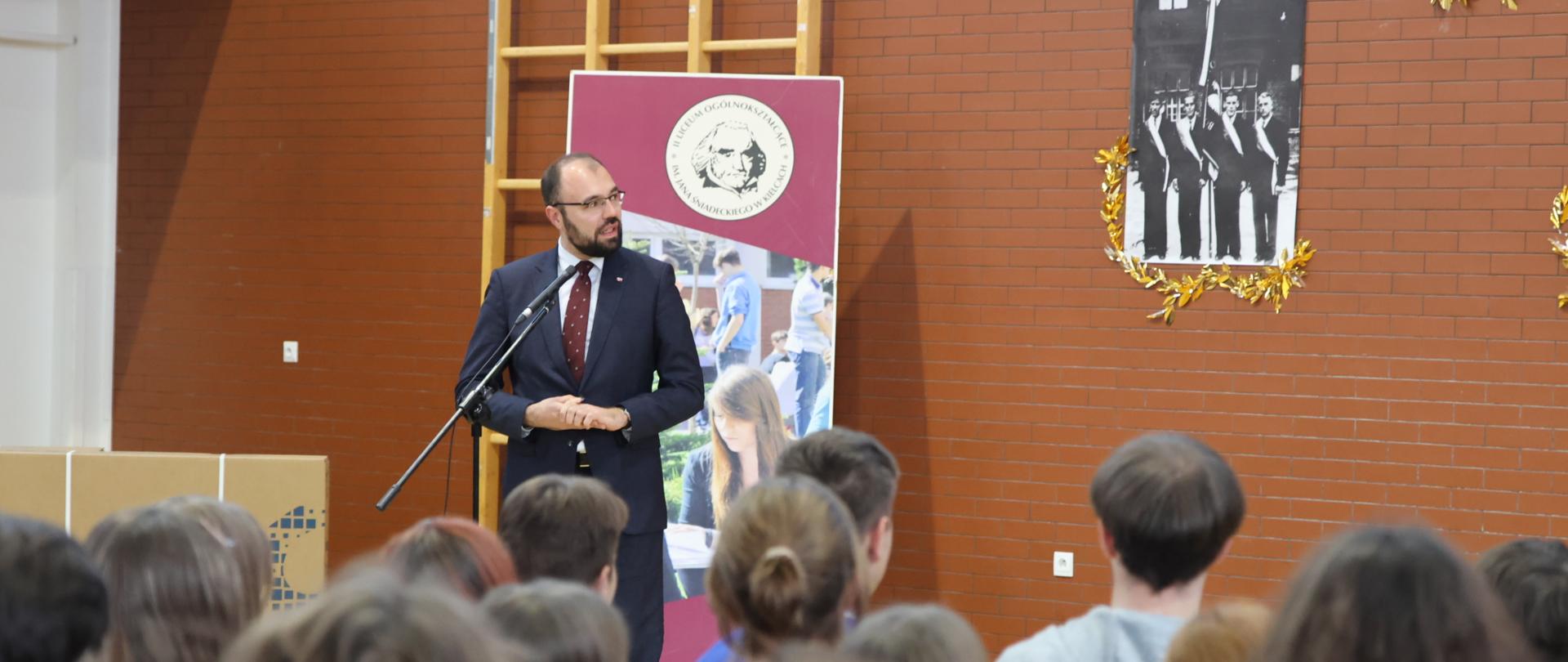 Na zdjęciu minister Szczucki stojący przy mikrofonie i przemawiający do zgromadzonych na sali, siedzących na krzesłach uczniów. W tle brązowa ściana, a na niej czarno-białe zdjęcie przedstawiające czterech mężczyzn ze sztandarem. Obok plansza dotycząca liceum.