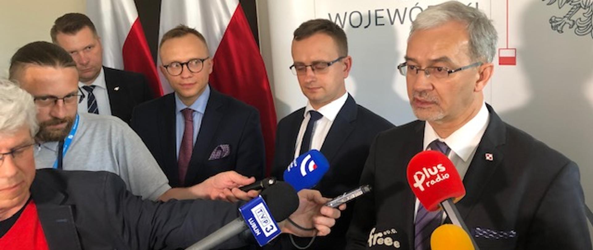 Dziennikarze z mikrofonami skierowanymi do ministra Jerzego Kwiecińskiego obok którego stoją wojewódzki inspektor nadzoru budowlanego, wiceminister Artur Soboń i wojewoda Przemysław Czarnek