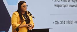 Minister funduszy i polityki regionalnej Małgorzata Jarosińska-Jedynak stoi przy pulpicie i przemawia do mikrofonu, za jej plecami grafika z prezentacją
