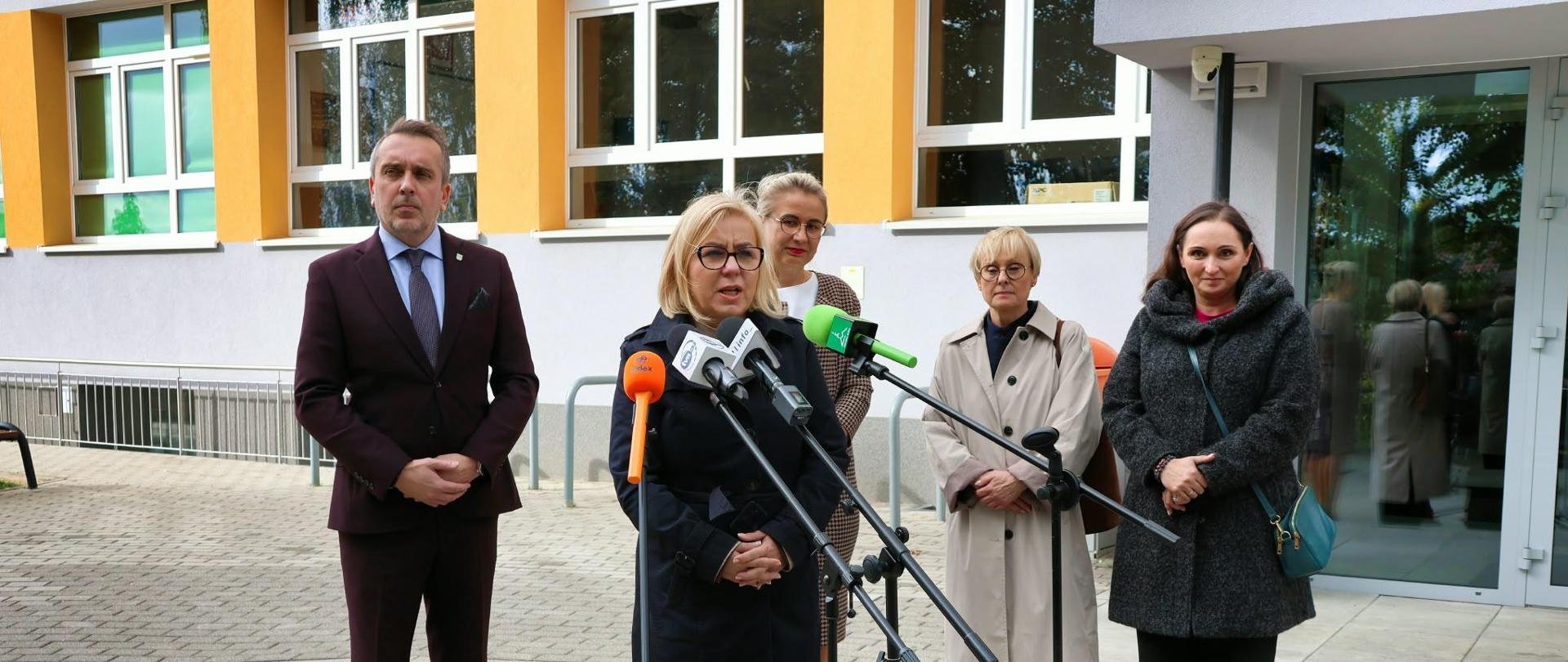 Ministra klimatu i środowiska Paulina Hennig-Kloska podczas konferencji prasowej w Szkole Podstawowej nr 14 w Zielonej Górze. Na zdjęciu ministra stoi przed budynkiem szkoły i mówi do mikrofonów dziennikarzy. Za nią stoi kilka osób.