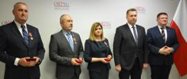Uczestnicy uroczystości stoją pod ścianą, obok nich polska flaga.