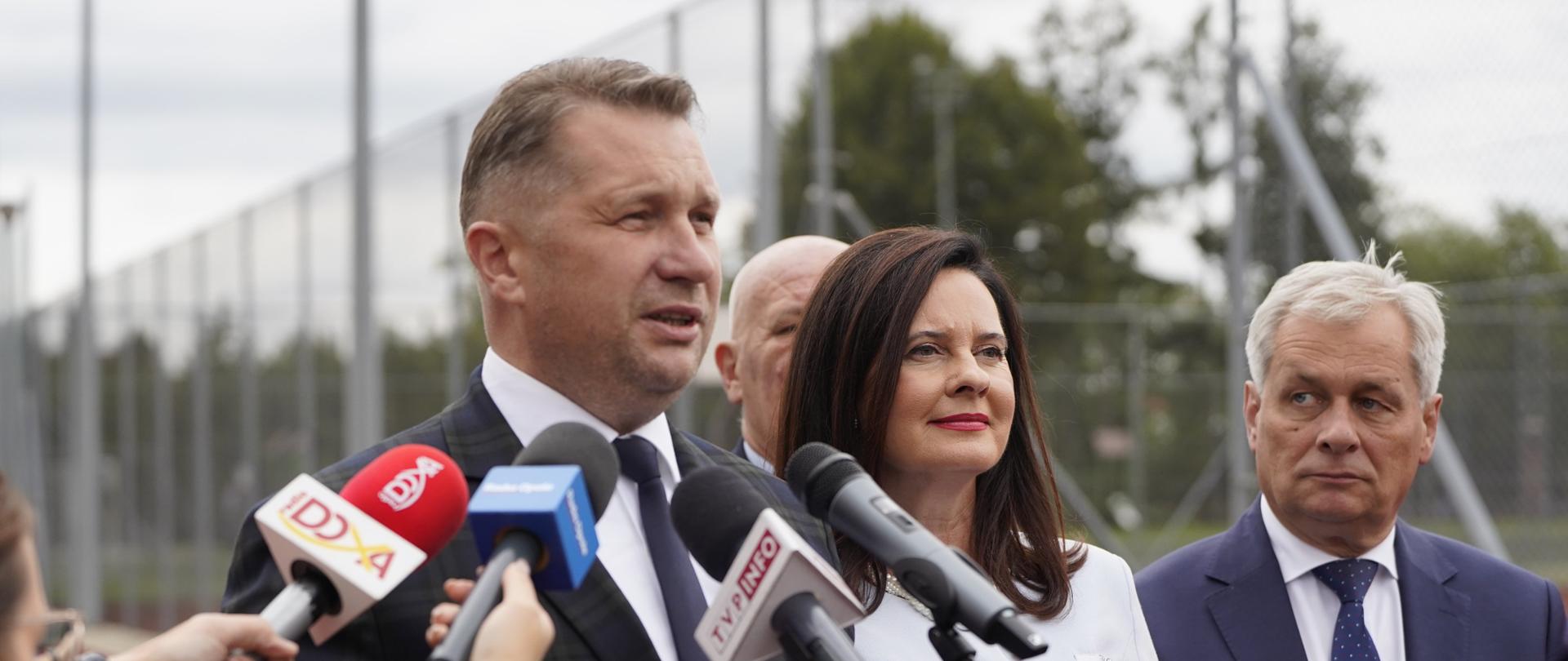 Na dworze otoczonym wysoką siatką boiskiem stoi minister Czarnek, trzyma w ręku broszurkę w brązowej okładce z napisem Demokracja bezpośrednia i mówi do mikrofonów, obok niego kobieta w białym ubraniu trzyma taką samą broszurkę, dalej mężczyzna w szarym garniturze.