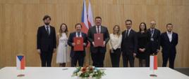Za stołem na którym stoją małe chorągiewki - flagi Polski i Czech stoi w rzędzie dziewięć osób, dwóch mężczyzn trzyma czerwone teczki.