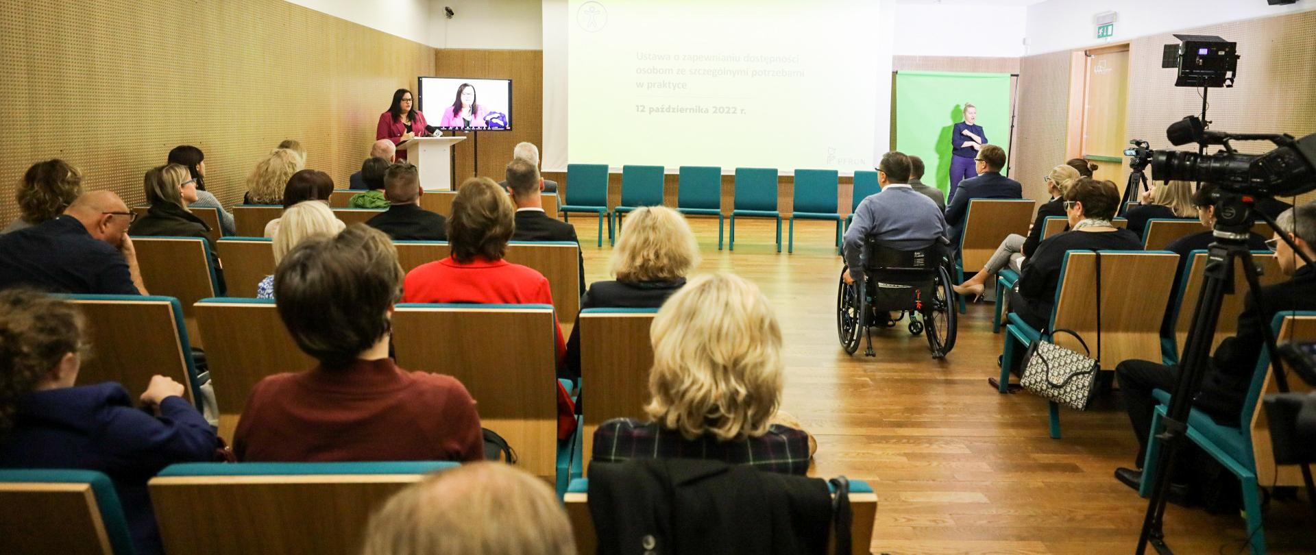 Grupa osób siedzi na krzesłach w sali konferencyjnej. Na końcu sali wiceminister Małgorzata Jarosińska-Jedynak w mównicy.