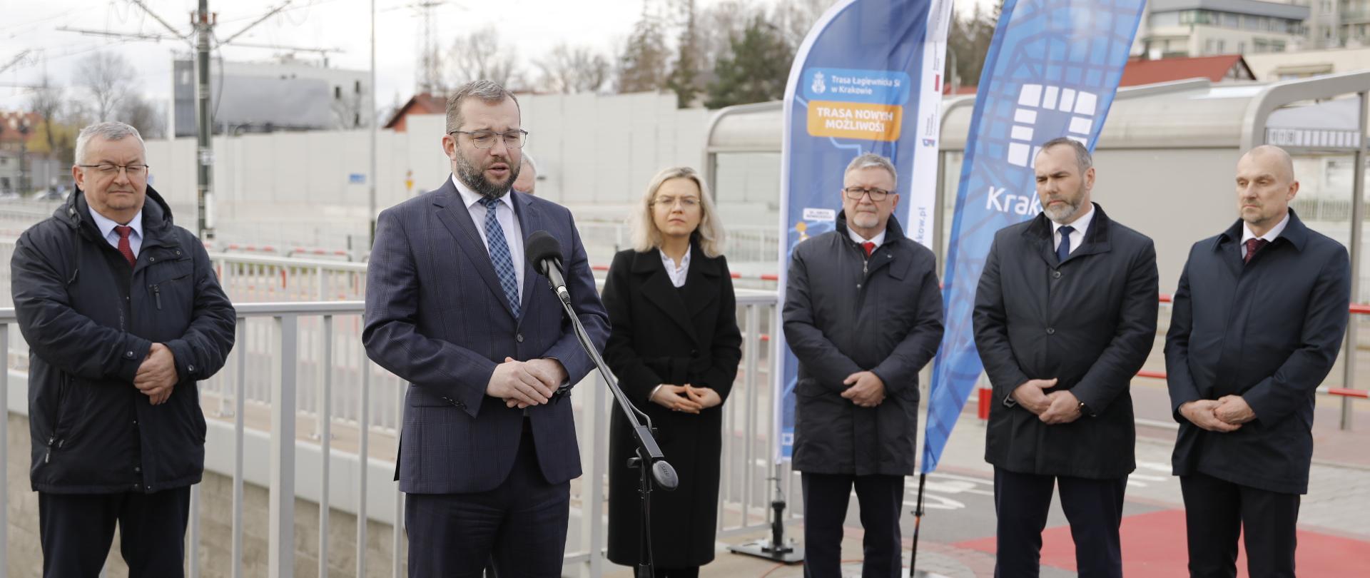 Minister Grzegorz Puda stoi i mówi do mikrofonu. W tle za jego plecami stoi sześć osób.
