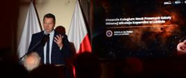 W mrocznej sali za mównicą stoi minister Czarnek i mówi do mikrofonu, za nim z boku wielki ekran z napisem Otwarcie Kolegium Nauk Prawnych Szkoły Głównej Mikołaja Kopernika.