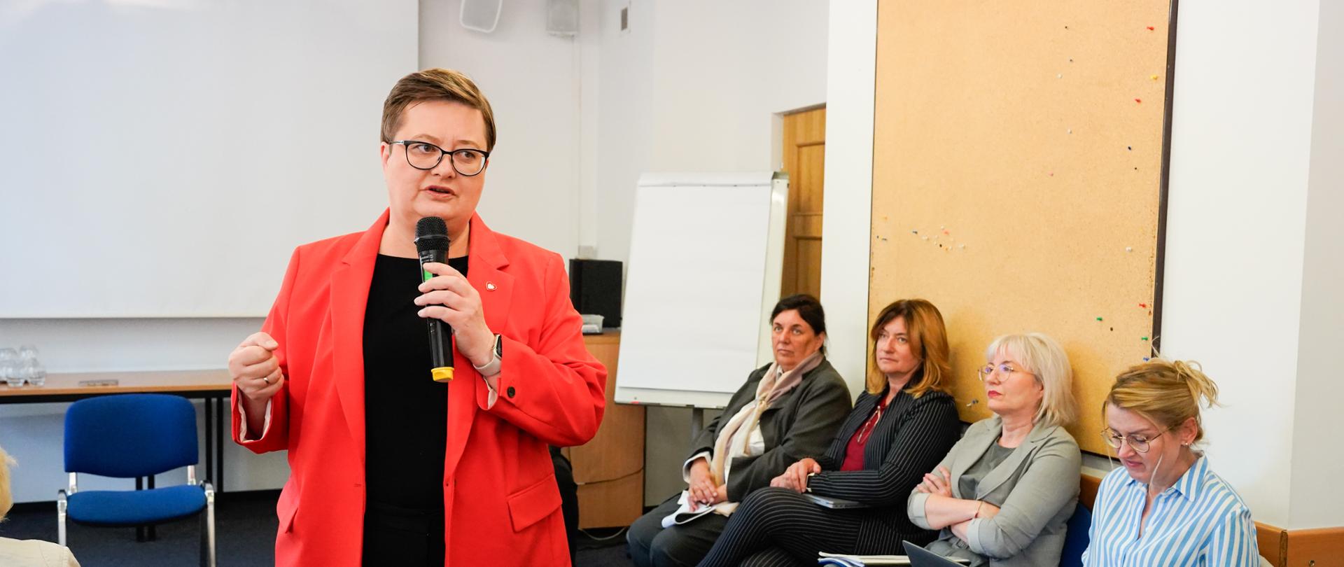 Kobieta w czerwonej marynarce i okularach przemawia do mikrofonu, gestykulując dłonią. Za nią siedzi kilka innych osób, a w tle widoczny jest biały ekran projekcyjny.