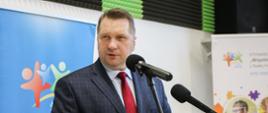Minister stoi i mówi do dwóch mikrofonów na stojakach, za nim kolorowe plakaty i ściana w czarno-zielony wzorek.