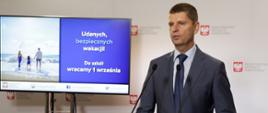 Minister Dariusz Piontkowski przemawia stojąc za mównicą, w tle prezentacja na monitorze "Udanych, bezpiecznych wakacji! Do szkół wracamy 1 września"