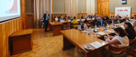 W sali ze ścianami wyłożonymi drewnianymi panelami przy ustawionych w czworokąt stołach siedzi dużo ludzi, w tle pod ścianą baner z napisem NAWA, z przodu na ścianie duży ekran, pod nim stoi mężczyzna w garniturze i mówi.
