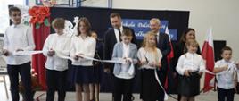 Pod ściana stoi w rzędzie siedmioro dzieci, trzymają nożyczki i przecinają białą rozciągniętą w poprzek przed nimi wstążkę, za nimi stoi minister Czarnek i mężczyzna w garniturze.