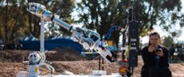 Na piaszczystym podłożu stoi robot z ramieniem, obok niego młoda kobieta, w tle drzewa.