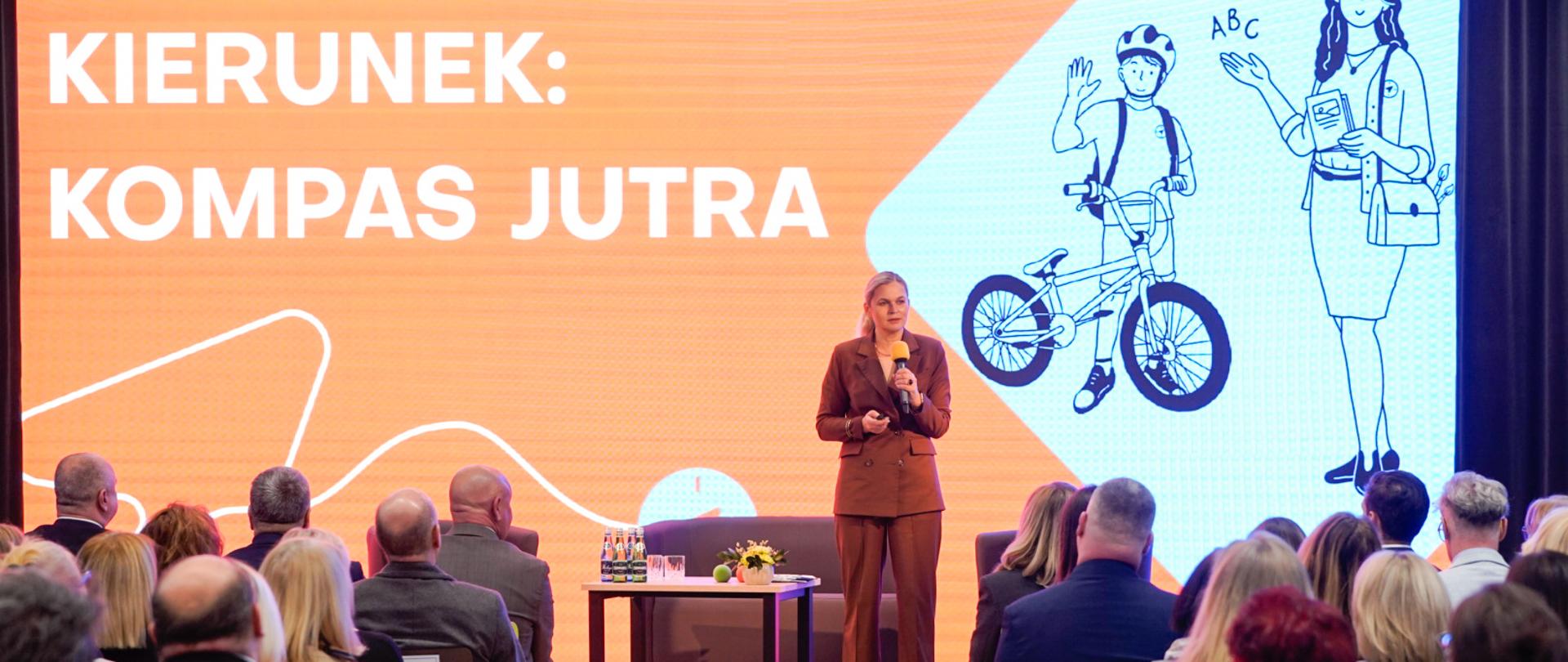 Na tle ekranu z napisem: "Kierunek: Kompas Jutra" stoi Minister Edukacji Barbara Nowacka. Minister Nowacka trzyma w lewej dłoni mikrofon, a obok niej znajduje się stolik z wodą, drugim mikrofonem i bukietem kwiatów. Minister Nowacka jest zwrócona do publiczności, którą są nauczyciele.