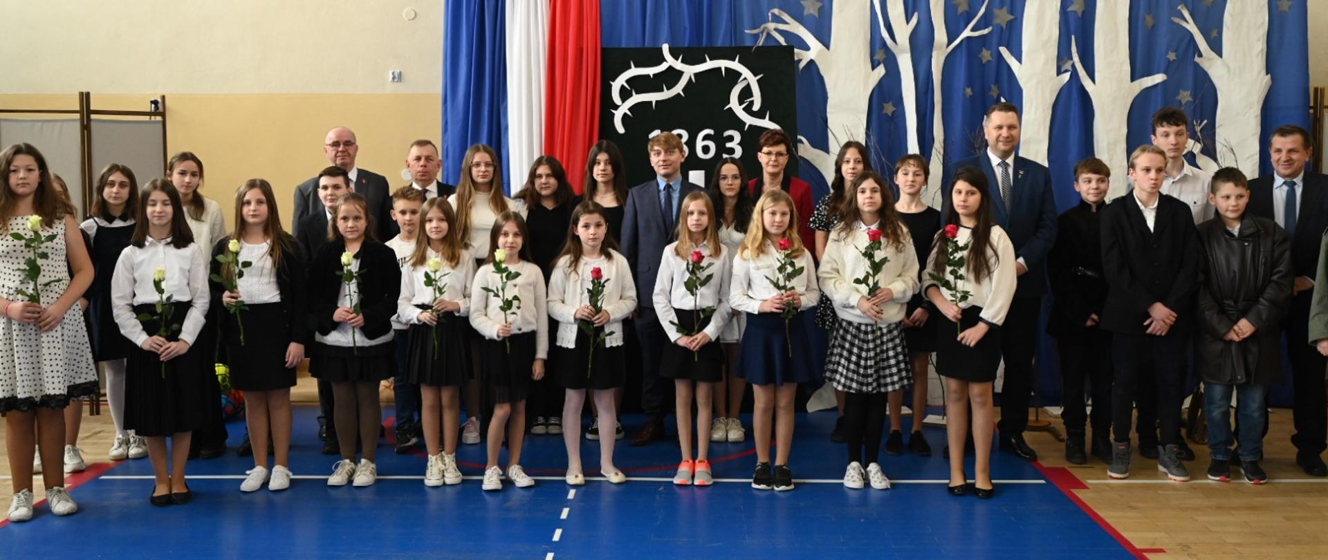 W sali gimnastycznej pod ścianą z koszem do koszykówki stoi grupa galowo ubranych dzieci, za nimi kilkoro dorosłych.