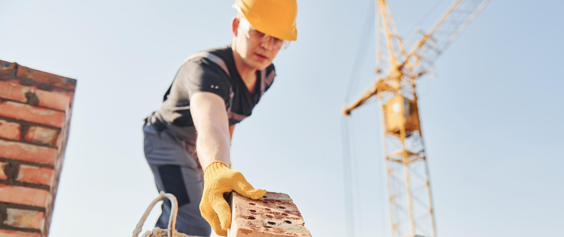 Holding brick and using hammer. Construction worker in uniform and safety equipment have job on building.