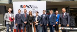 Na dworze w słońcu stoi w szeregu grupa elegancko ubranych osób, mężczyzna po lewej trzyma bukiet kwiatów, za nimi ścianka z napisem SOWA, dalej wejście do budynku.