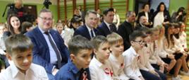Szkolna sala gimnastyczna z drabinkami przy ścianach, na ustawionych w rzędy krzesłach siedzą elegancko ubrane dzieci i kilkoro dorosłych, wśród nich minister Czarnek.