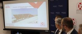 Minister Grzegorz Puda siedzi przy stole na konferencji w Szczyrku i ogląda wyświetlaną na ekranie prezentację nt. rozbudowy infrastruktury sportowej na Kubalonce