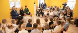 W sali z drewnianą podłogą i żółtymi ścianami na krzesłach siedzą minister Czarnek i dwie kobiety, wokół nich na ustawionych w półkole krzesełkach siedzą dzieci.