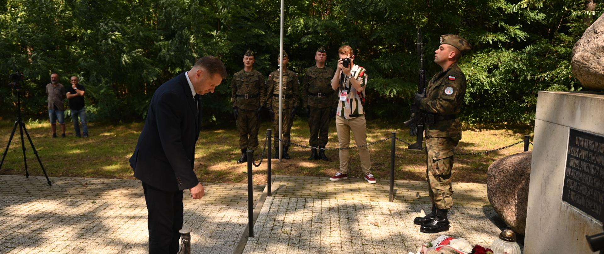 Minister Czarnek pochyla się przed pomnikiem - kamieniem na postumencie, przed którym leża biało-czerwone kwiaty.