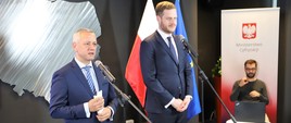 Minister cyfryzacji Marek Zagórski i wiceminister zdrowia Janusz Cieszyński przemawiają do mikrofonów. Za nimi siedzi tłumacz PJM. 