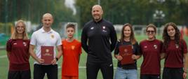 Zdjęcie. Minister sportu i turystyki Jakub Rutnicki stoi na boisku piłkarskim razem z przedstawicielami i zawodnikami klubu UKS Varsovia oraz oraz Akademii Piłkarska Diamonds Academy.