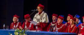 Przy niebieskim stole siedzą ludzie w czerwonych ceremonialnych strojach, kobieta stoi i mówi do mikrofonu.