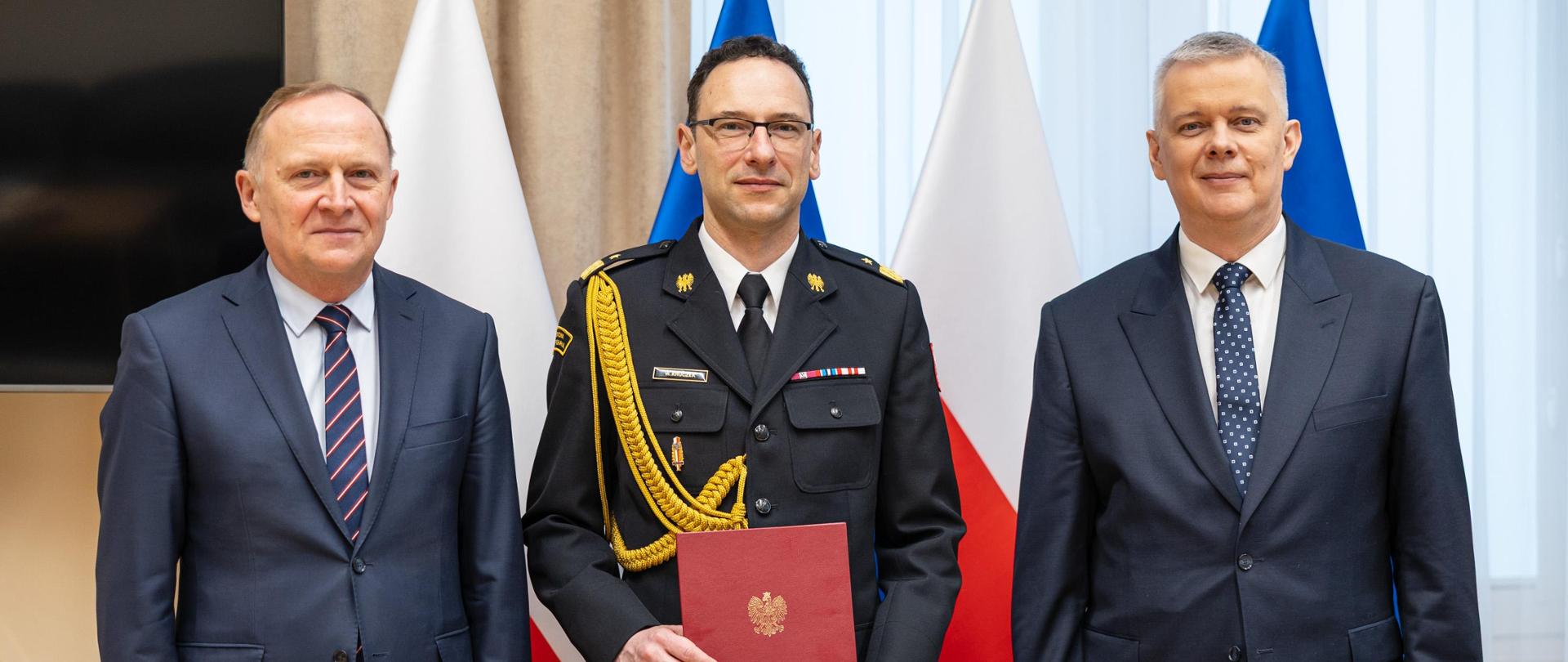 Uroczyste wręczenie nominacji strażakowi w obecności urzędników na tle flag państwowych.