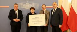 Pod szarą ścianką z napisem Śląski Urząd Wojewódzki w Katowicach stoi trzech mężczyzn w garniturach, wśród nich kobieta w czarnym ubraniu trzyma białą tabliczkę z napisem Branżowe Centrum Umiejętności.