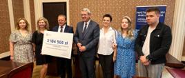 Przed ścianą stoi minister Murdzek i grupa ludzi, jeden z mężczyzn trzyma duży symboliczny czek z napisem 2 184 500 zł.