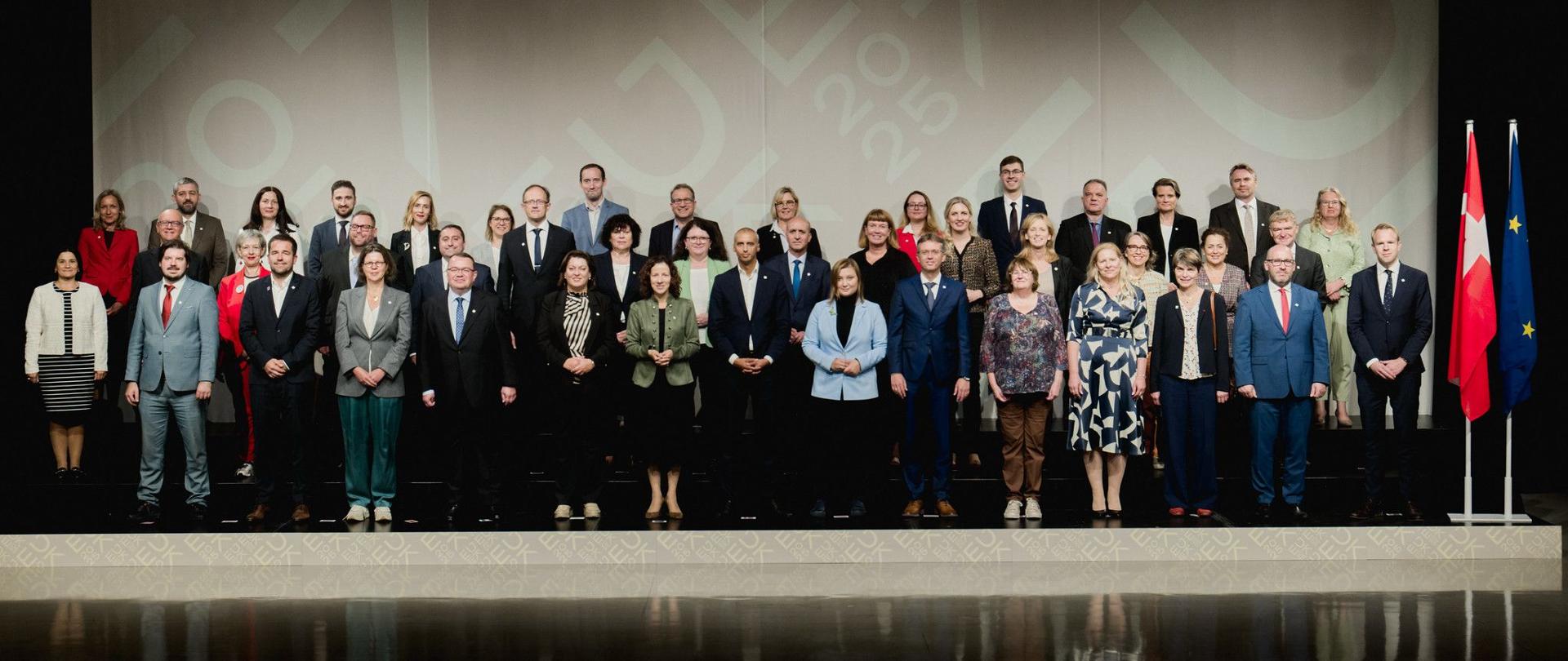 Duża grupa osób, ubranych w stroje formalne i biznesowe, stoi w kilku rzędach na scenie w pomieszczeniu o nowoczesnym wystroju. W tle znajduje się baner z niewyraźnymi napisami, a po prawej stronie stoją dwie flagi. Wszystkie osoby patrzą prosto w obiektyw, a ich odbicie jest widoczne na błyszczącej podłodze.