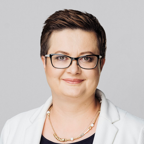 Na tle jasnoszarej ściany stoi wiceminister Katarzyna Lubnauer w białej marynarce.