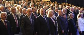 Widok z przodu pod kątem na wielka aulę wypełnioną stojącymi ludźmi.
