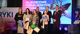 Grupa młodych osób stoi na scenie. Za nimi trzech mężczyzn w garniturach. W tle ekran z wyświetlanym tekstem: Młodzieżowe Forum Nowych Technologii
