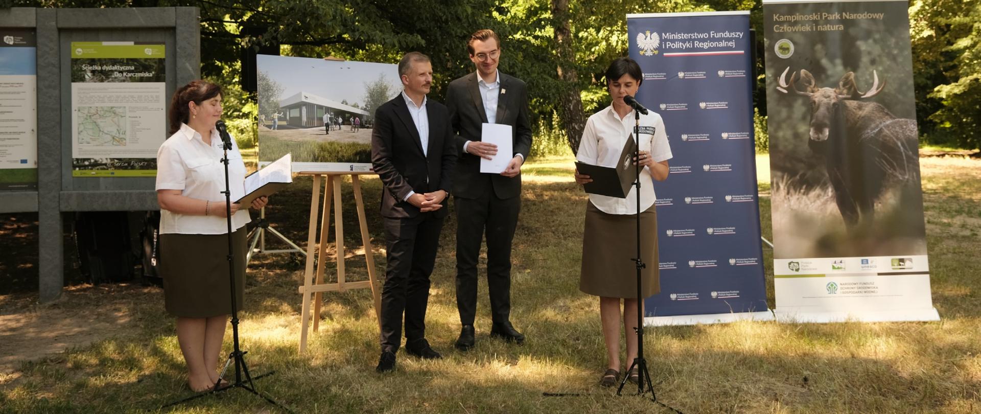 Cztery osoby stoją w lesie przed mikrofonami. Za nimi banery. Wśród osób wiceminister Jan Szyszko.