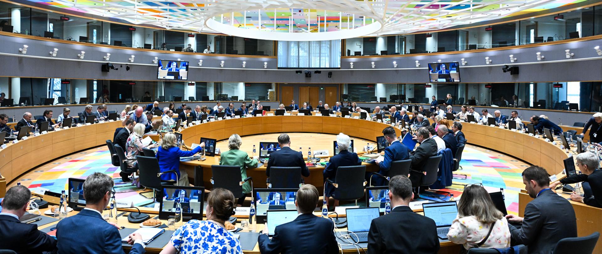 Uczestnicy posiedzenia Rady ECOFIN siedzą przy okrągłym stole konferencyjnym w dużej sali z kolorowym sufitem i ekranami.