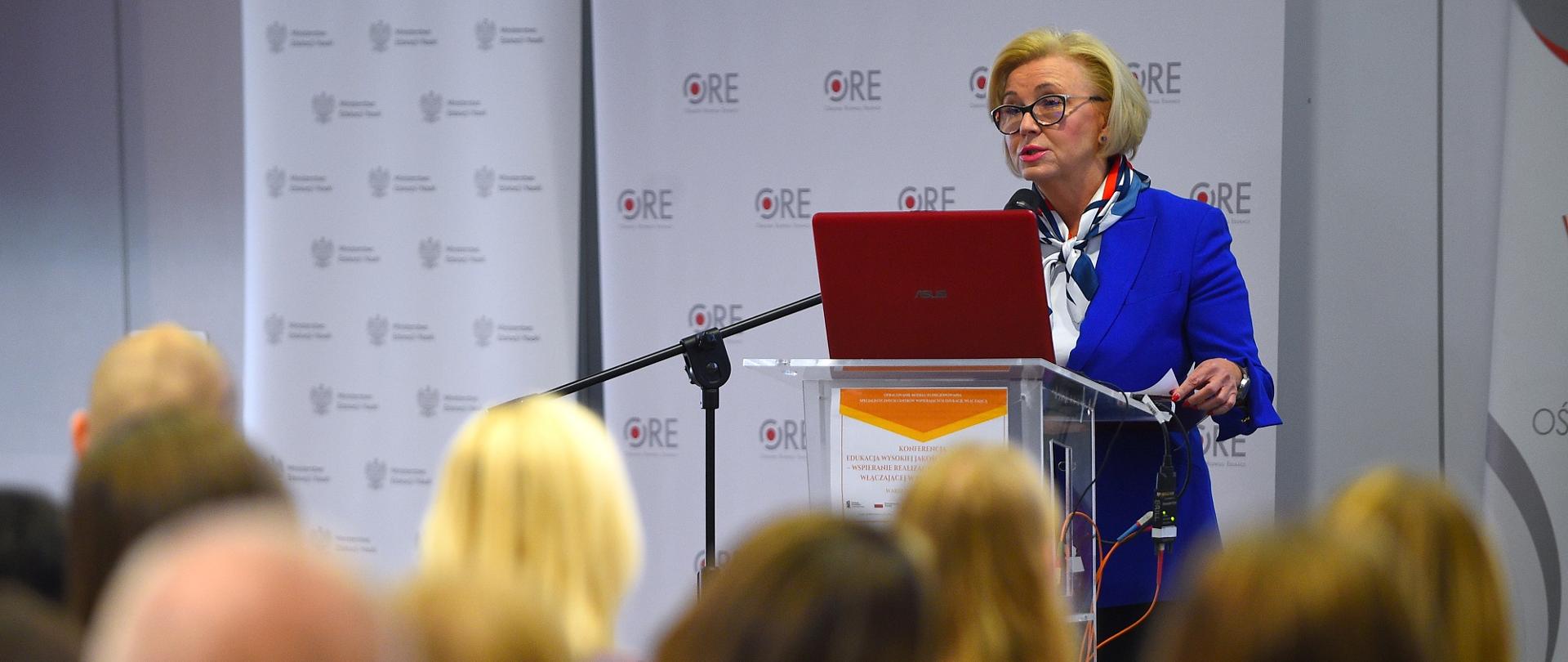 Minister Machałek w niebieskim ubraniu stoi i mówi do mikrofonu, za nią ściana z napisami ORE.