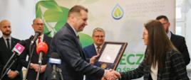 Minister Czarnek stoi i ściska rękę kobiecie w marynarce w czarno-żółtą kratkę, przed nimi kilka mikrofonów na stojakach, za nimi grupa mężczyzn w garniturach, jeden z nich trzyma dyplom w ramkach.