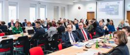 Wiele osób siedzi na sali przy długich stołach. W tle na ścianie logo FEPW 2021-2027.