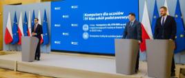 Na podwyższeniu przy szarych mównicach stoi trzech mężczyzn w garniturach, za nimi niebieska ściana z napisem Komputery dla uczniów IV klas szkół podstawowych.