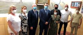 Minister Przemysław Czarnek w otoczeniu nauczycieli ze Szkoły Podstawowej w Warcie. Na twarzach wszyscy mają maseczki. W tle widoczny fragment obrazu oraz gablota. Pomarańczowa posadzka z czarno-białymi elementami. 