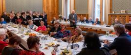 Duża sala ze ścianami w zielony wzorek, przy drewnianych stołach ustawionych w czworokąt siedzi dużo elegancko ubranych osób. Pod ścianą stoi chór.