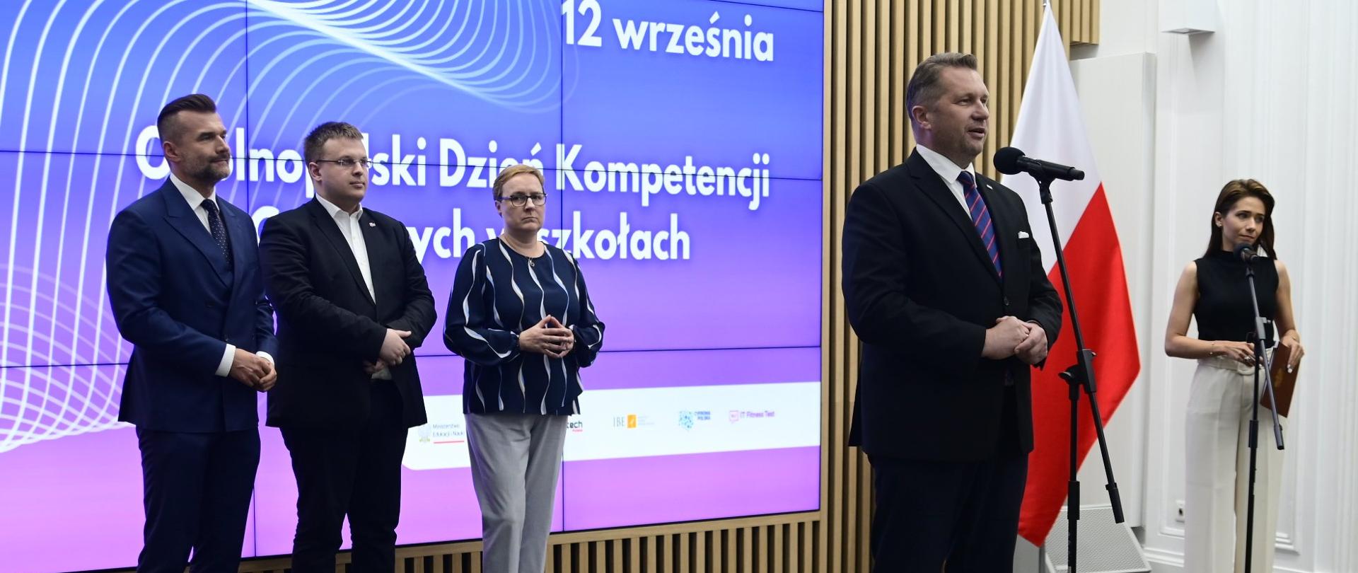 W sali stoi minister Czarnek i mówi do mikrofonu na stojaku, za nim z boku kobieta stoi przy drugim mikrofonie, dalej pod ścianą z wielkim ekranem wyświetlającym napis 12 września - ogólnopolski dzień kompetencji cyfrowych w szkołach stoi dwóch mężczyzn i kobieta czarnej bluzie w białe paski.