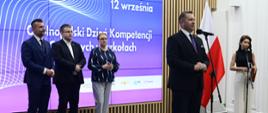 W sali stoi minister Czarnek i mówi do mikrofonu na stojaku, za nim z boku kobieta stoi przy drugim mikrofonie, dalej pod ścianą z wielkim ekranem wyświetlającym napis 12 września - ogólnopolski dzień kompetencji cyfrowych w szkołach stoi dwóch mężczyzn i kobieta czarnej bluzie w białe paski.