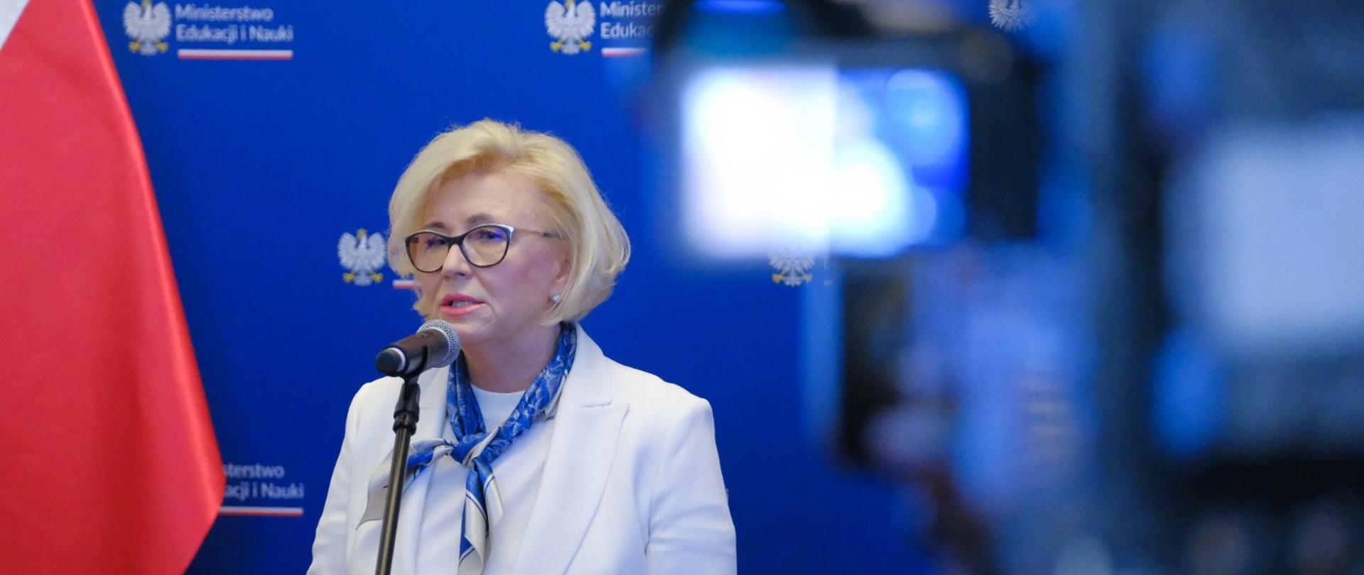Wiceminister Machałek stoi i mówi do mikrofonu na stojaku, za nią niebieska ścianka z napisem Ministerstwo Edukacji i Nauki, obok polska flaga, przed nią kamera.