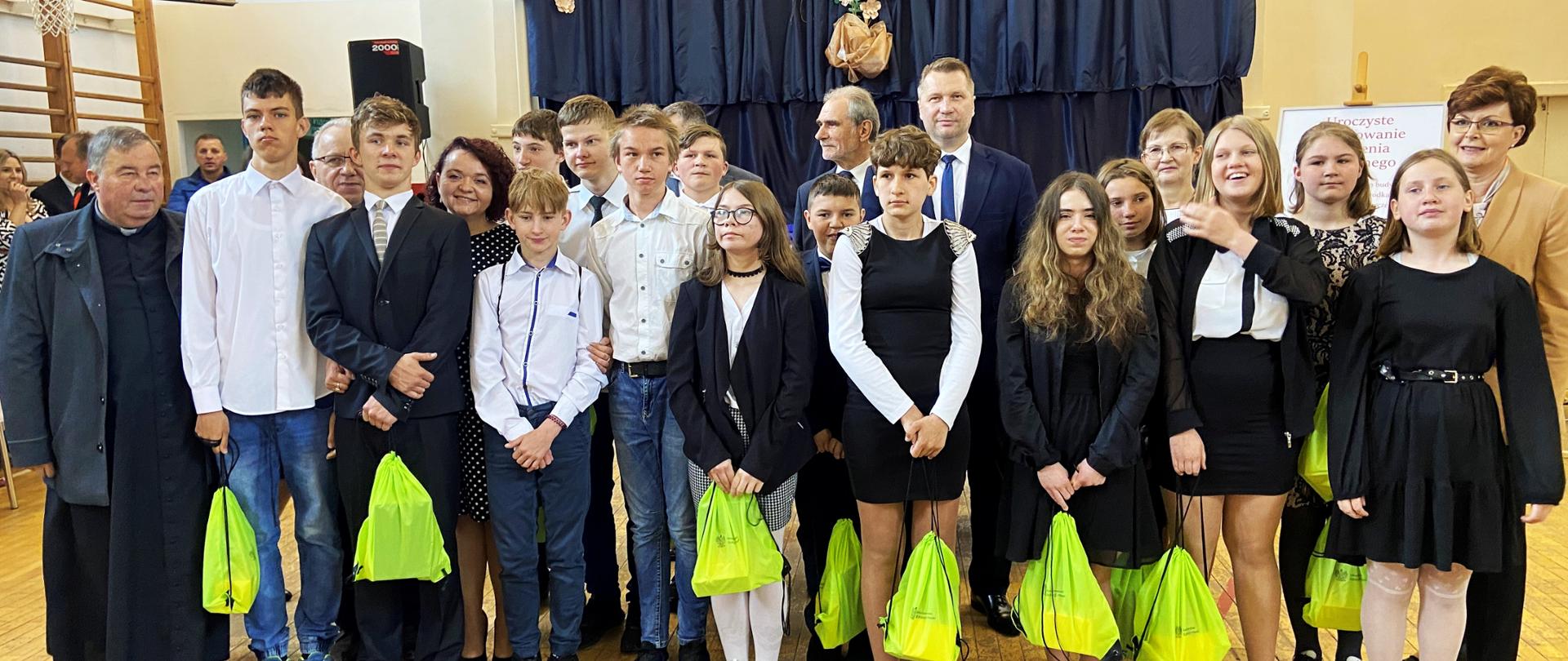 Na sali gimnastycznej stoi grupa ludzi, z przodu ubrane na galowo dzieci trzymające jaskrawozielone worki, za nimi minister Czarnek i kilka osób, za nimi w tle ściana przysłonięta granatową kotarą.
