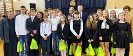 Na sali gimnastycznej stoi grupa ludzi, z przodu ubrane na galowo dzieci trzymające jaskrawozielone worki, za nimi minister Czarnek i kilka osób, za nimi w tle ściana przysłonięta granatową kotarą.